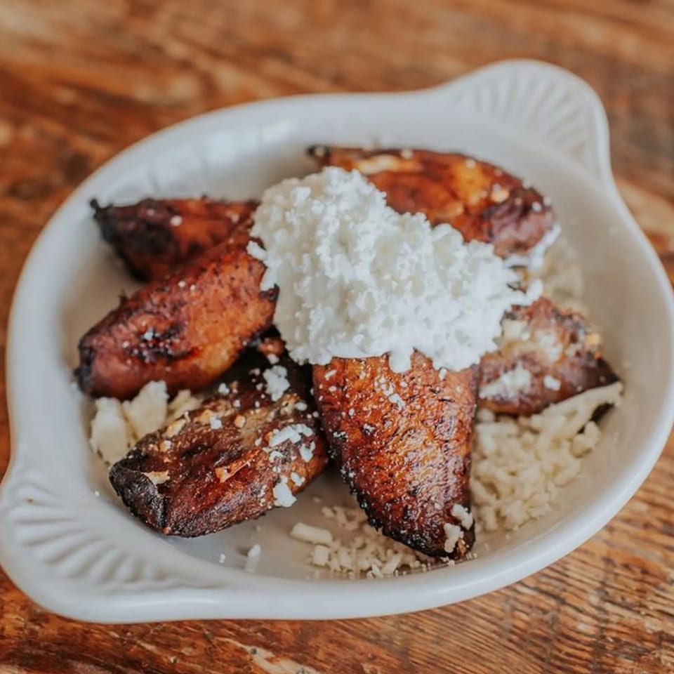 Sweet plantains topped with white cheese and cream, served in a white dish on a wooden table.