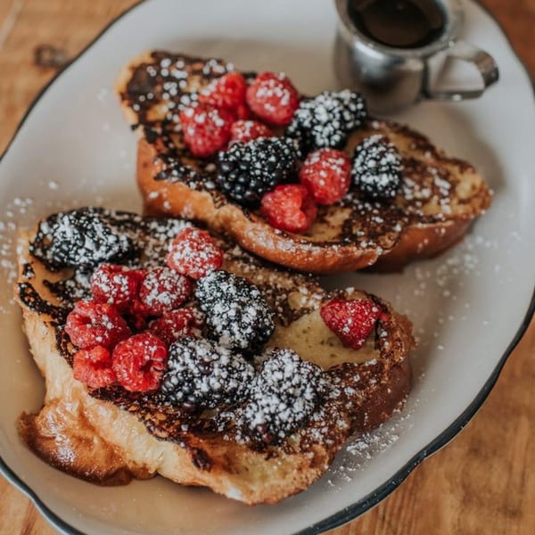 Two slices of French toast topped with raspberries, blackberries, and powdered sugar, with syrup.