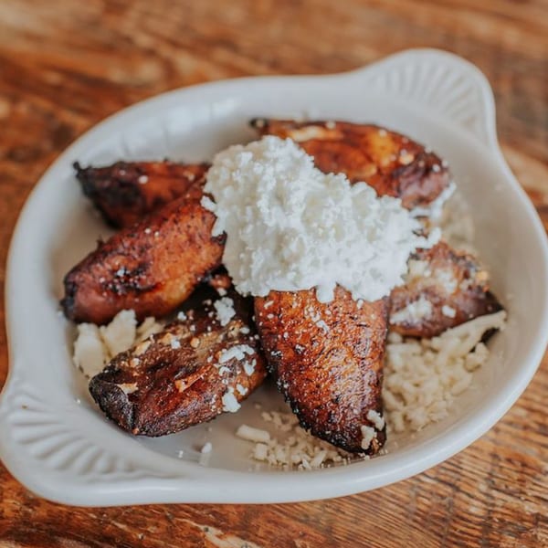 Fried plantains topped with white cheese and cream, served in a white dish on a wooden table.