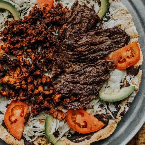 Mexican tlayuda topped with grilled steak, chorizo, shredded cabbage, tomato, and avocado slices.