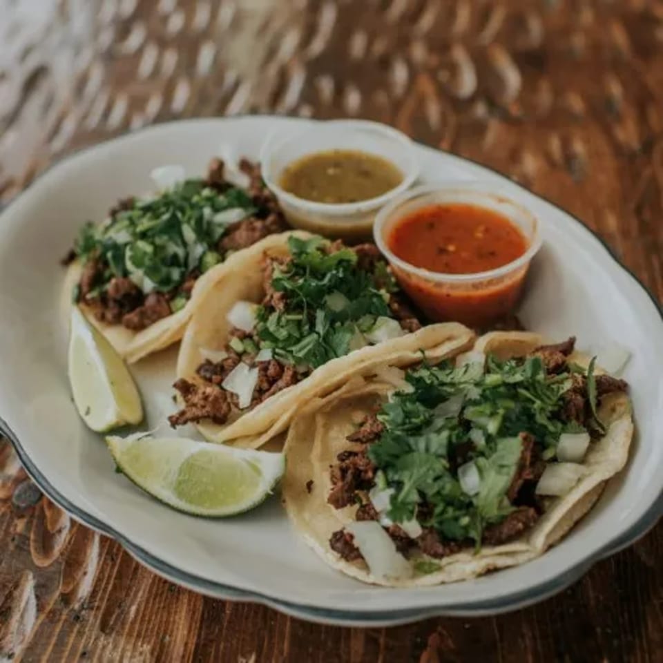 Three Mexican street tacos with meat, cilantro, onions, lime wedges, and two salsa cups.