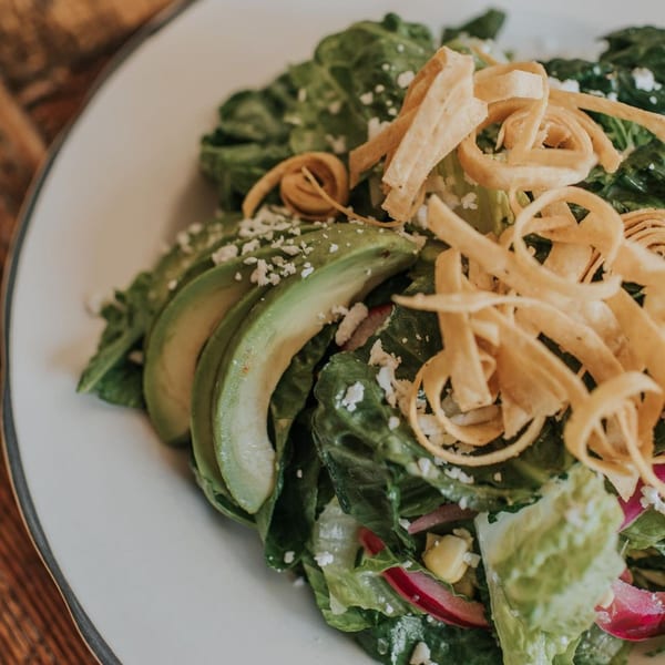 Mexican salad with lettuce, avocado slices, tortilla strips, and crumbled cheese on a white plate.