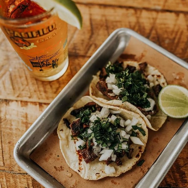 Two carne asada tacos with onion and cilantro, a lime wedge, and a beer on a wooden table.