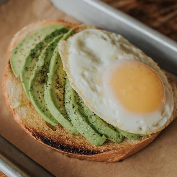 Avocado toast with sliced avocado and a fried egg, seasoned with pepper, on a brown surface.