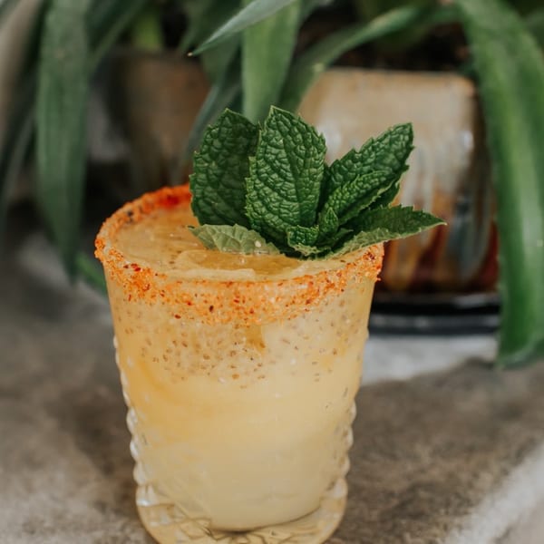 A refreshing yellow cocktail with a chili-salted rim and a mint garnish, set against a green plant background.